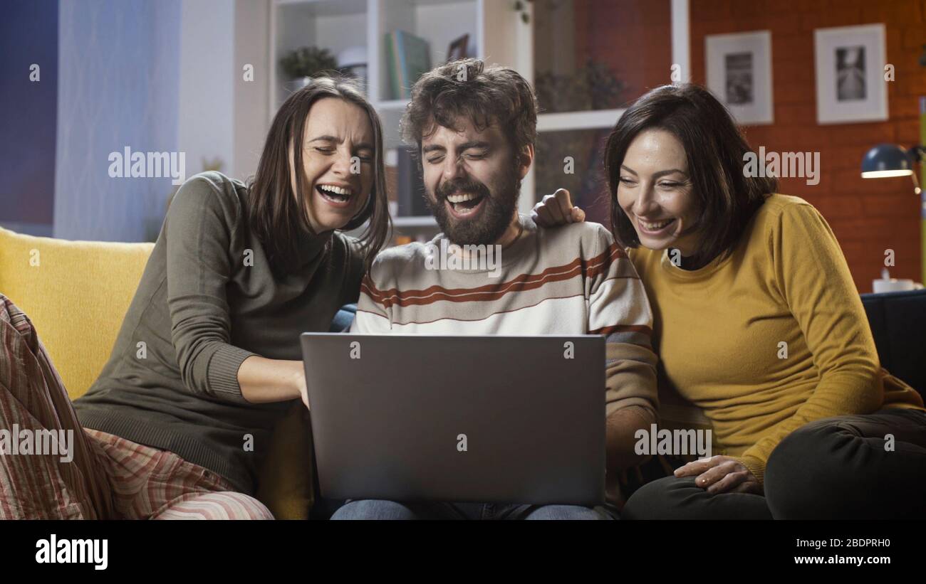 Des amis joyeux assis sur le canapé à la maison et se connectant en ligne avec leur ordinateur portable, ils sont des réseaux sociaux et s'amuser Banque D'Images