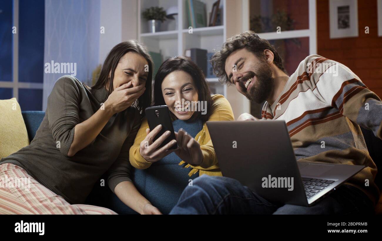 Les amis assis sur le canapé à la maison et les réseaux sociaux avec un smartphone, ils rient et regardent des vidéos ensemble Banque D'Images