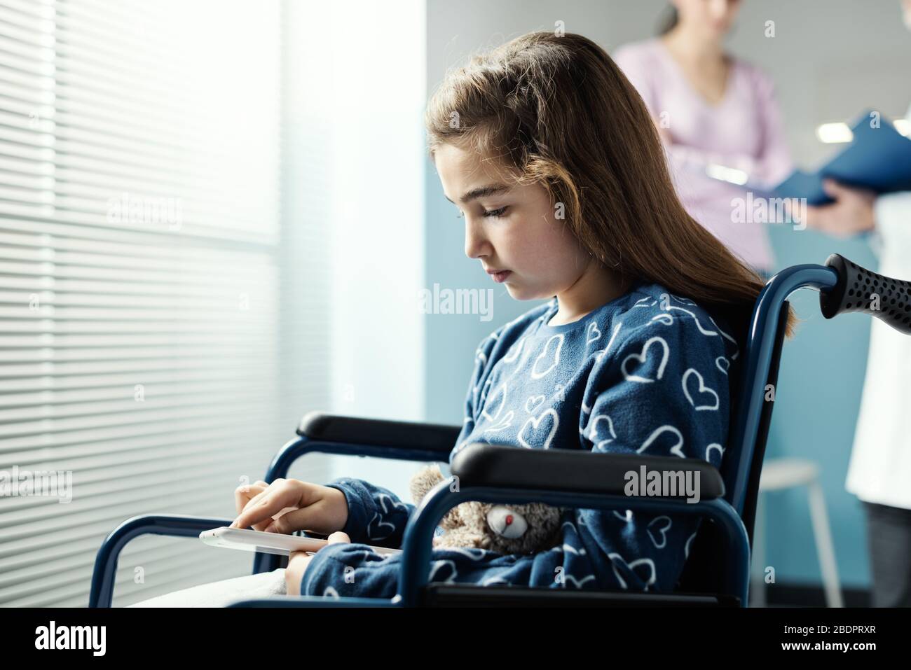 Jolie fille en fauteuil roulant à l'hôpital en utilisant un comprimé, sa mère et le médecin parlent en arrière-plan Banque D'Images