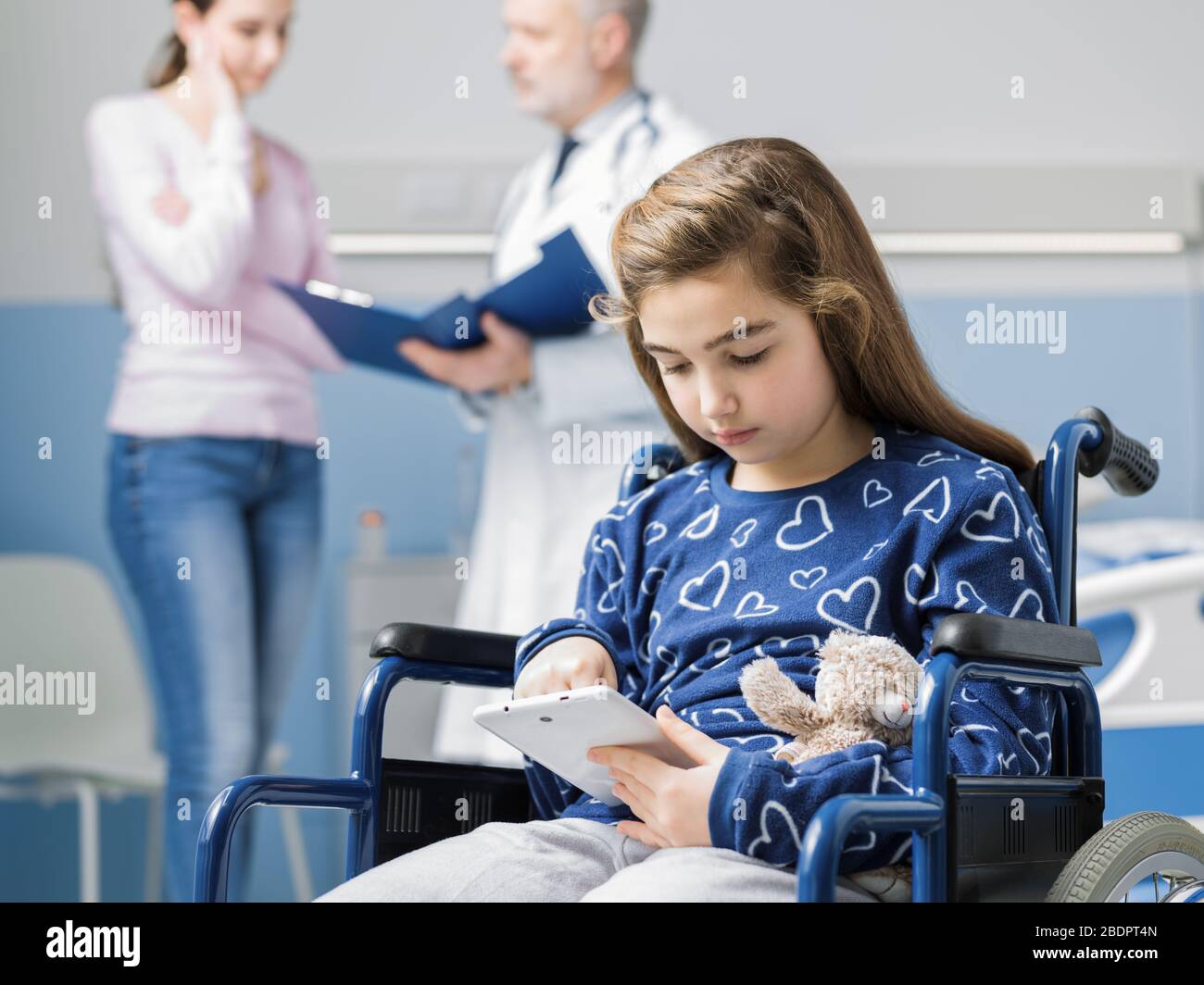 Fille en fauteuil roulant utilisant une tablette numérique à l'hôpital, sa mère et le médecin discutent en arrière-plan Banque D'Images