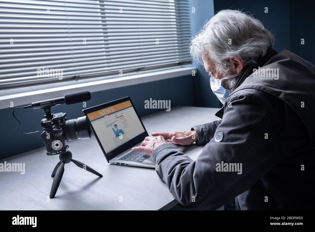 Homme senior assis au bureau et portant un masque chirurgical, il se connecte à son ordinateur portable et diffuse une vidéo en ligne sur son canal, covid-19 virus o Banque D'Images