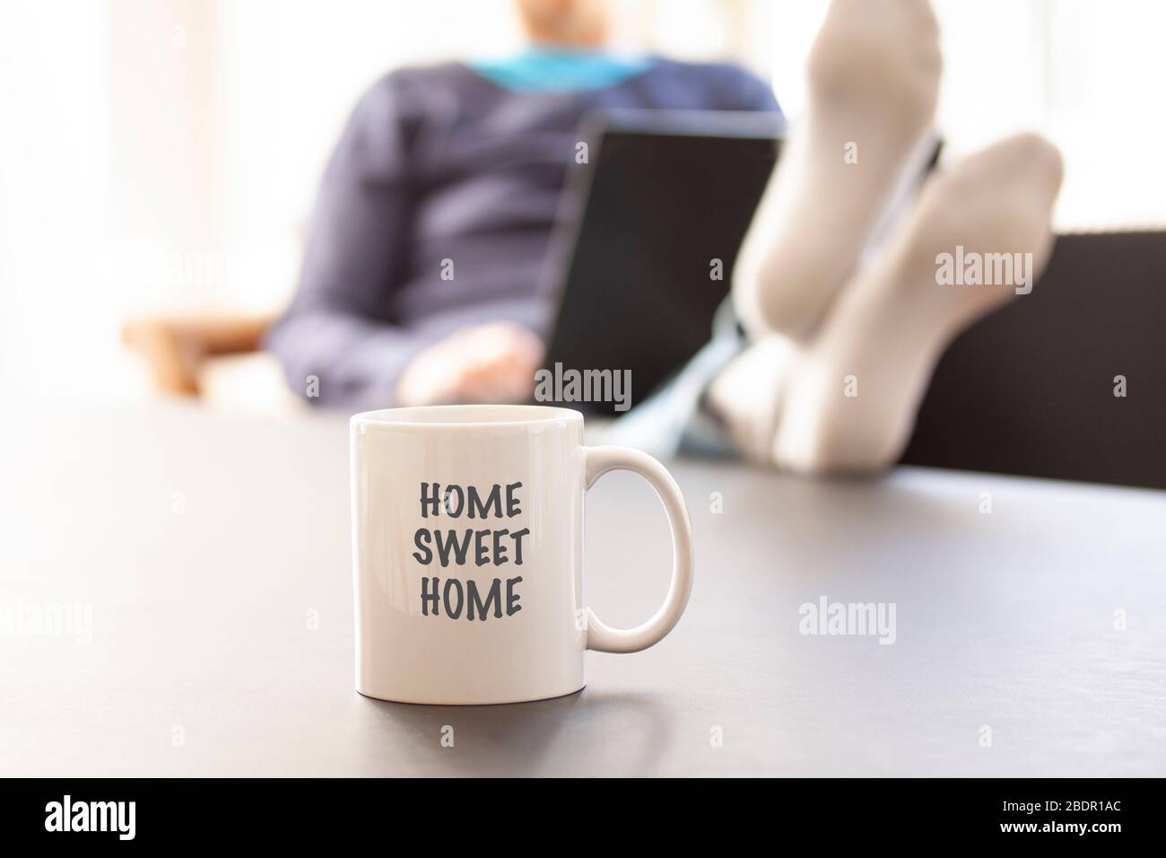 Femme assise dans un canapé avec un ordinateur portable une elle pieds sur une table où une tasse ou une tasse avec le texte ACCUEIL SWEET HOME est placé. Mise au point sélective Banque D'Images