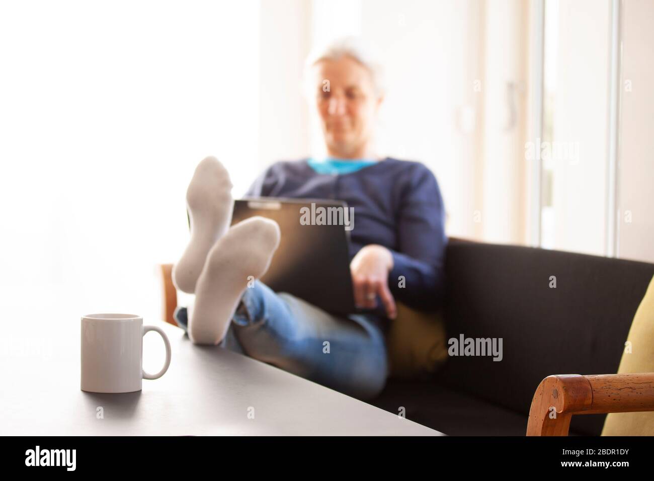 Femme assise dans un canapé avec un ordinateur portable et ses pieds sur la table où une tasse ou une tasse avec le texte ACCUEIL SWEET HOME est placé. De travail Banque D'Images