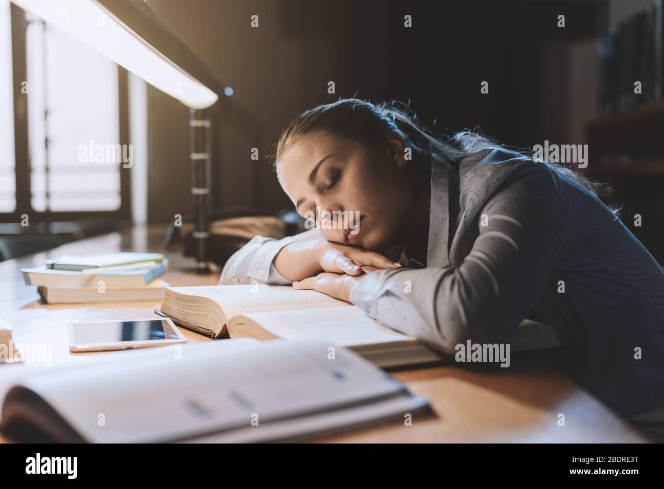 Étudiante féminine étudiant à la bibliothèque tard dans la nuit, elle se penche sur un livre ouvert et le sommeil, l'épuisement et le concept de surtravail Banque D'Images