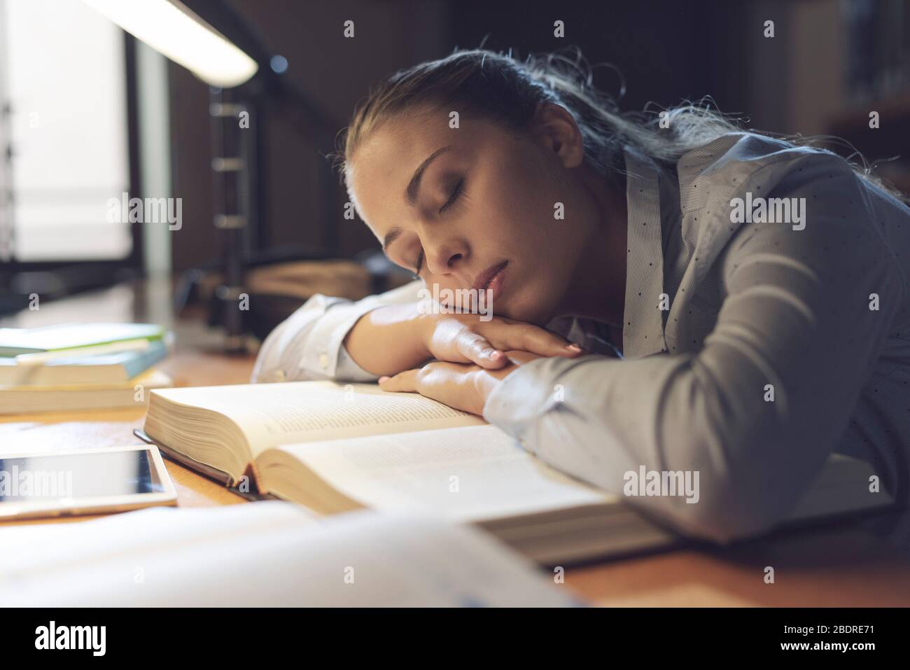 Étudiante féminine étudiant à la bibliothèque tard dans la nuit, elle se penche sur un livre ouvert et le sommeil, l'épuisement et le concept de surtravail Banque D'Images
