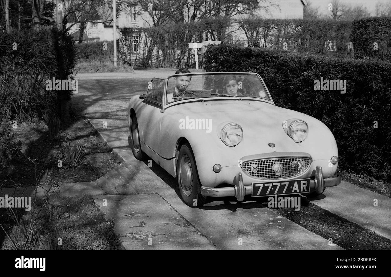 1960, historique, un couple assis dans une voiture de sport Austin Sprite garée à côté d'une haie sur une allée en béton étroit, Eastholm Green, Angleterre, Royaume-Uni. Banque D'Images
