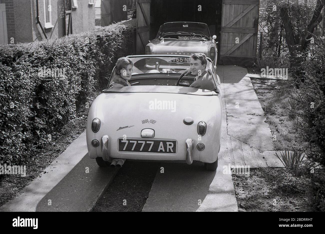 1960, historique, un jeune couple adulte assis dans une voiture de sport Austin Sprite garée à côté d'une haie sur une allée en béton étroit, Eastholm Green, Angleterre, Royaume-Uni. Une cicatrice de sport alpin Sunbeam se trouve également sur le trajet, garé à moitié à l'intérieur d'un garage en bois encadré. Banque D'Images