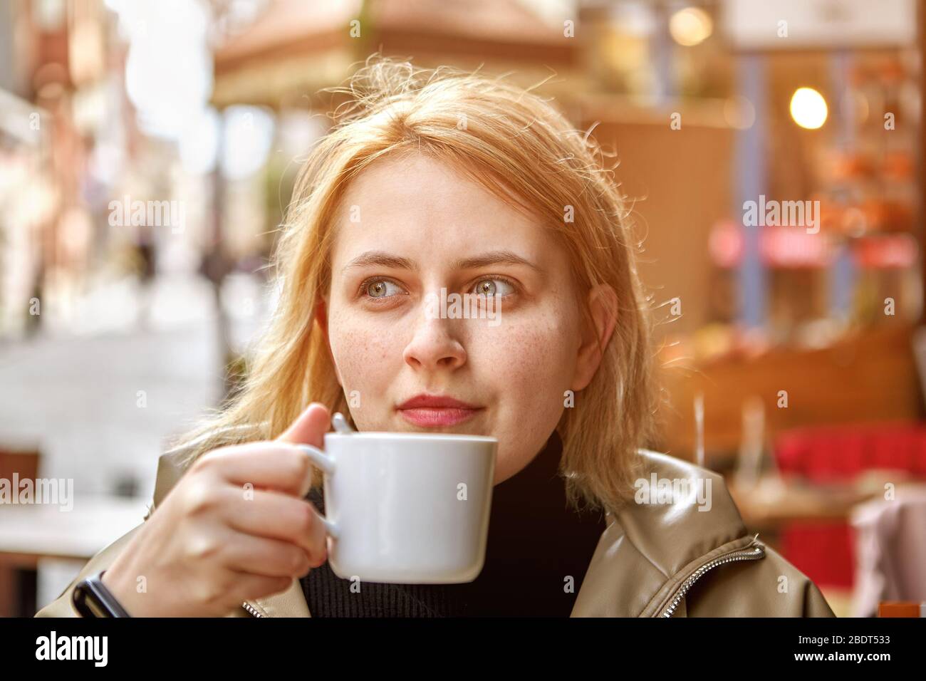 Une jeune femme blanche caucasienne aux cheveux blonds est assise dans le café extérieur avec une petite tasse de café dans ses mains. Banque D'Images