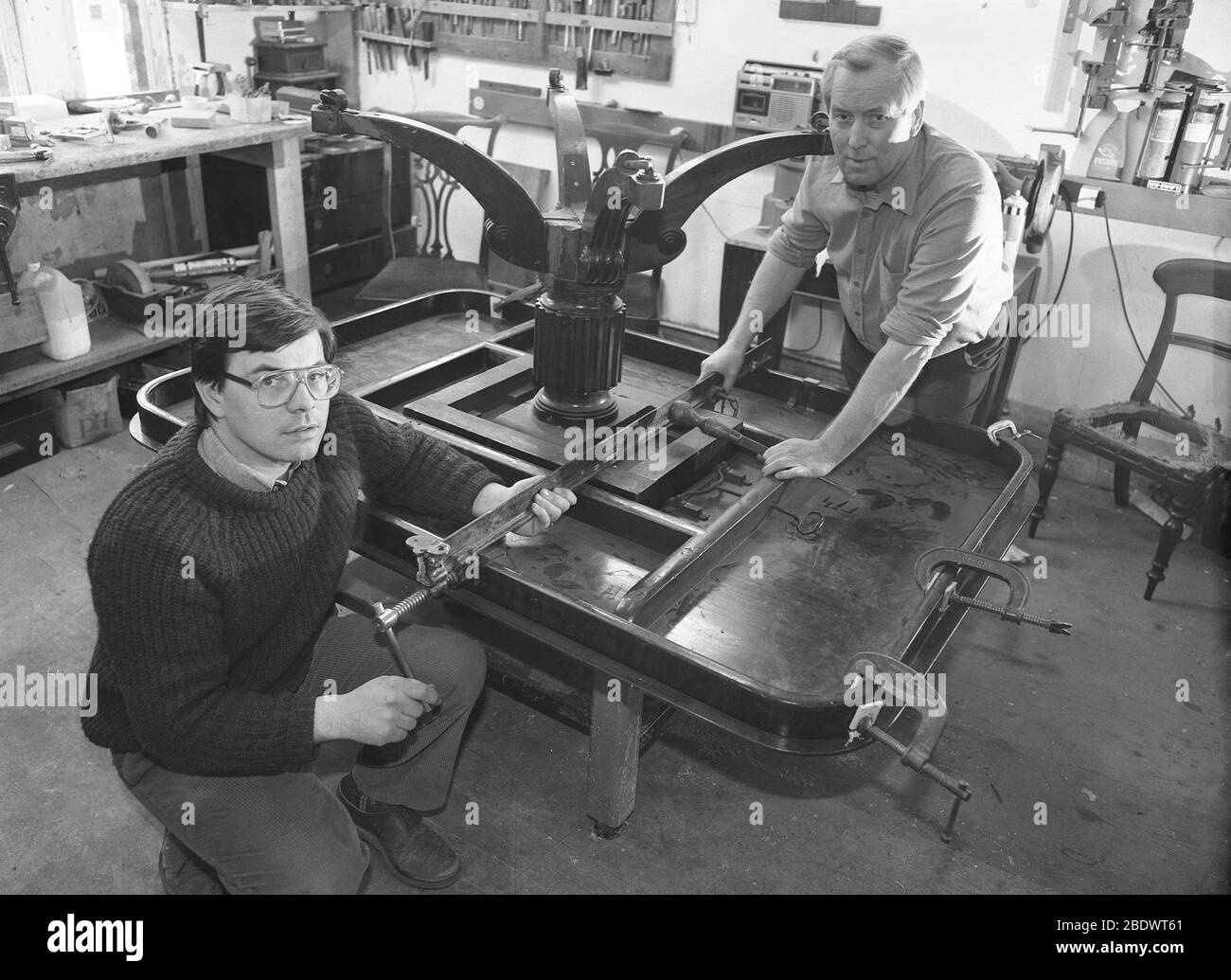 1980, deux restaurateurs de meubles de sexe masculin dans leur petit atelier, avec une table en bois antique, accroché à une autre table, pour faciliter l'accès au cadre et aux jambes, Angleterre, Royaume-Uni. Banque D'Images