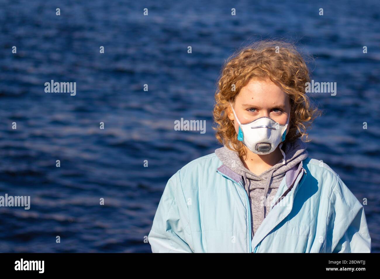Une fille dans un masque de protection médicale sur son visage regarde l'appareil photo avec un fond d'eau bleue. Femme avec cheveux corbiels blond. Espace de copie Banque D'Images
