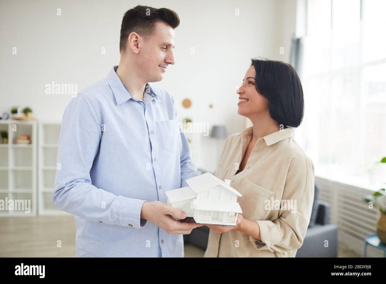 Jeune couple tenant le modèle de la maison et souriant les uns aux autres tout en se tenant dans le salon à la maison Banque D'Images