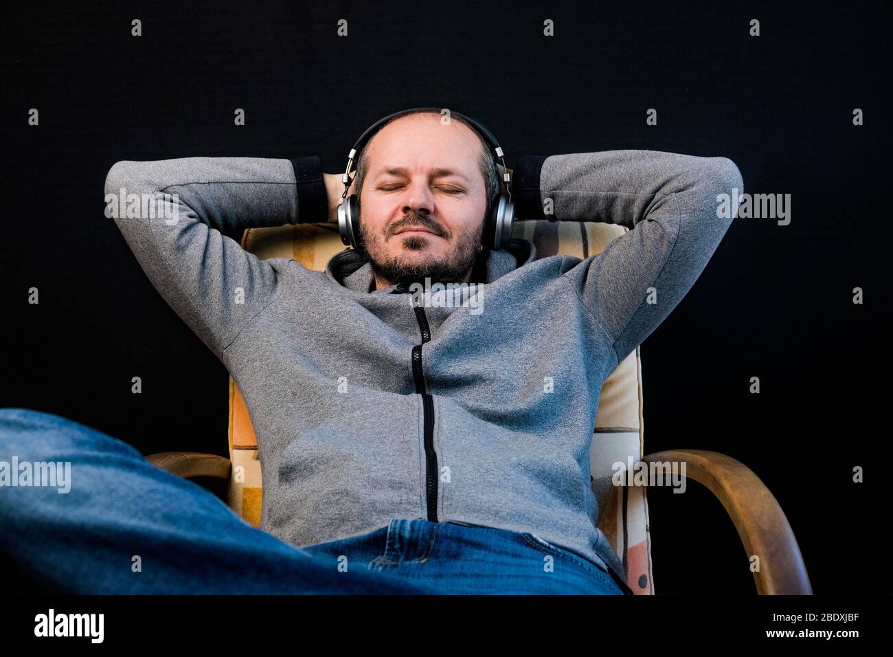 Homme barbu dans des vêtements décontractés hoodie assis dans une chaise avec casque, écoute de la musique Banque D'Images