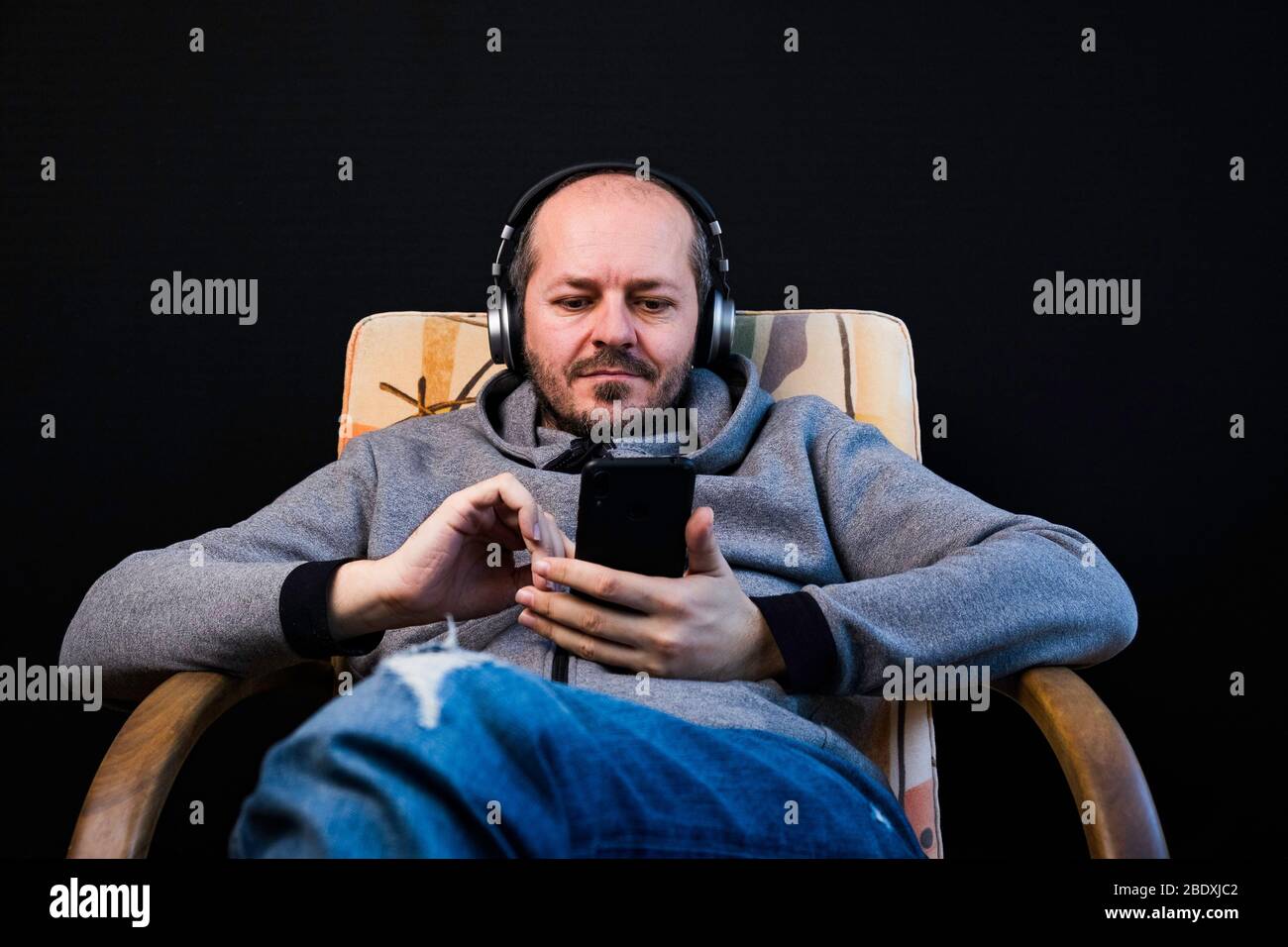 Homme barbu dans des vêtements décontractés hoodie assis dans une chaise avec casque, écoute de la musique Banque D'Images