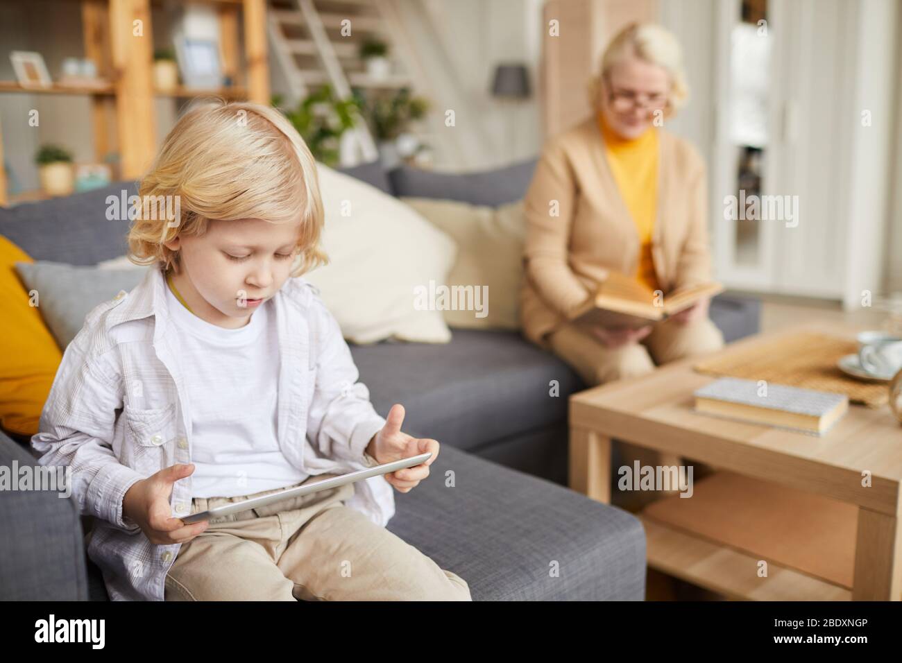 Petit garçon avec cheveux blond jouant jeu d'ordinateur sur tablette pc avec sa grand-mère assis sur un canapé en arrière-plan à la maison Banque D'Images
