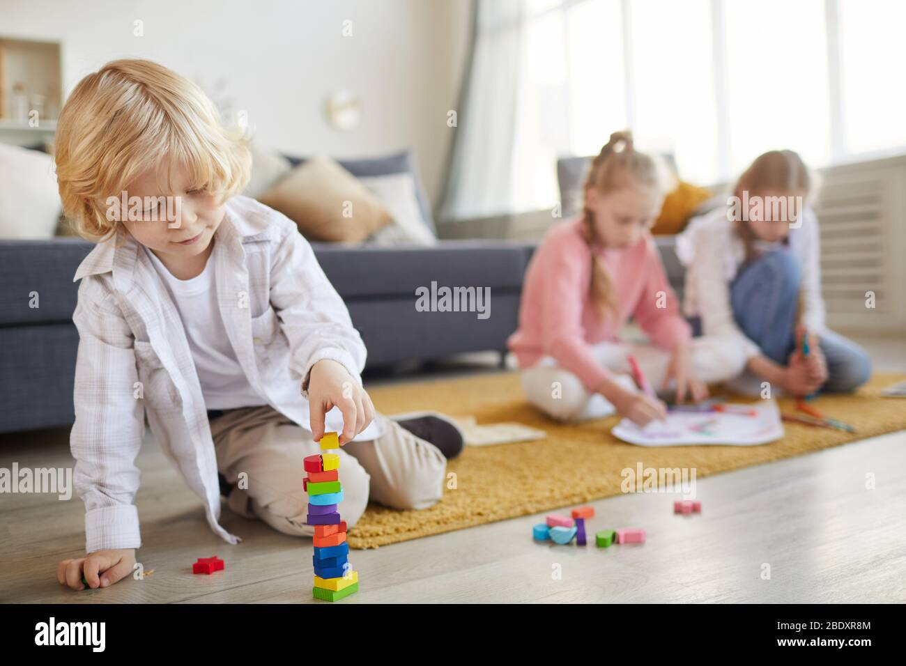 Petit garçon avec cheveux blond jouant sur le sol avec des jouets avec ses deux sœurs dessinant dans l'arrière-plan dans le salon Banque D'Images