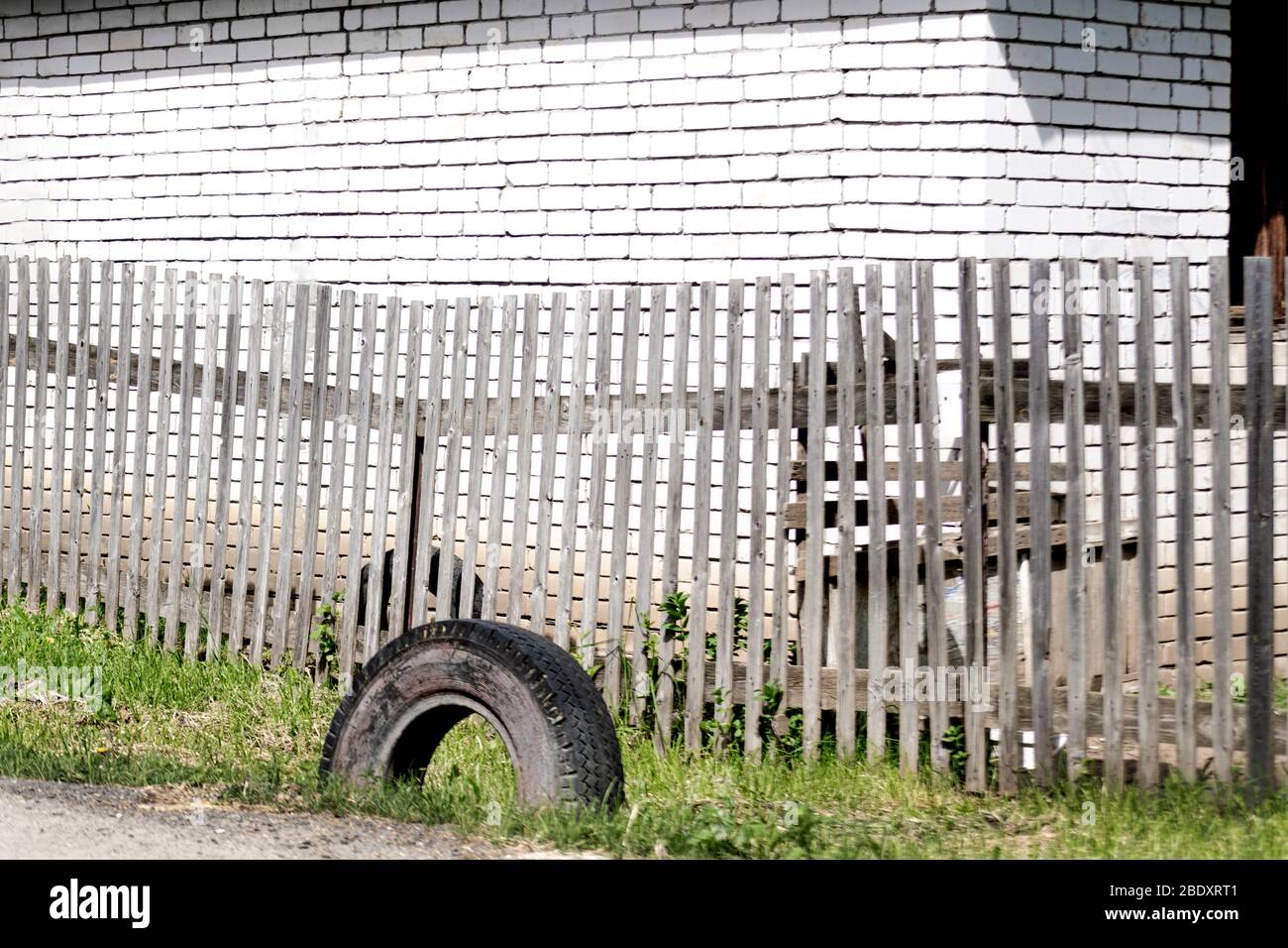 Une vieille clôture en bois gris de promenade avec des planches parallèles et un pneu usagé sur l'herbe verte devant un mur de brique blanche, lors d'une journée ensoleillée et brillante. A con Banque D'Images