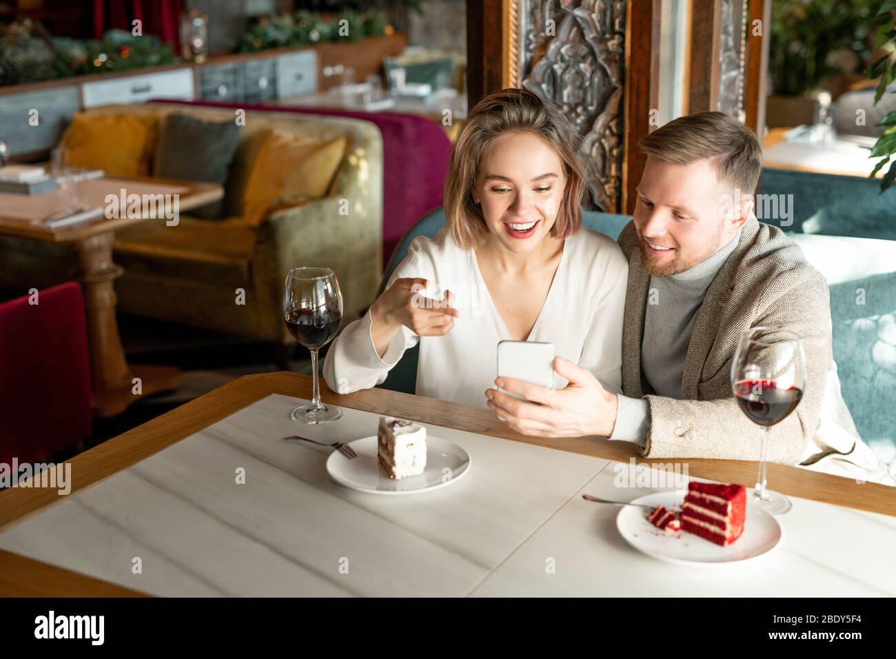Une jeune femme heureuse pointant sur l'écran du smartphone tout en discutant avec son petit ami par table servie au restaurant Banque D'Images