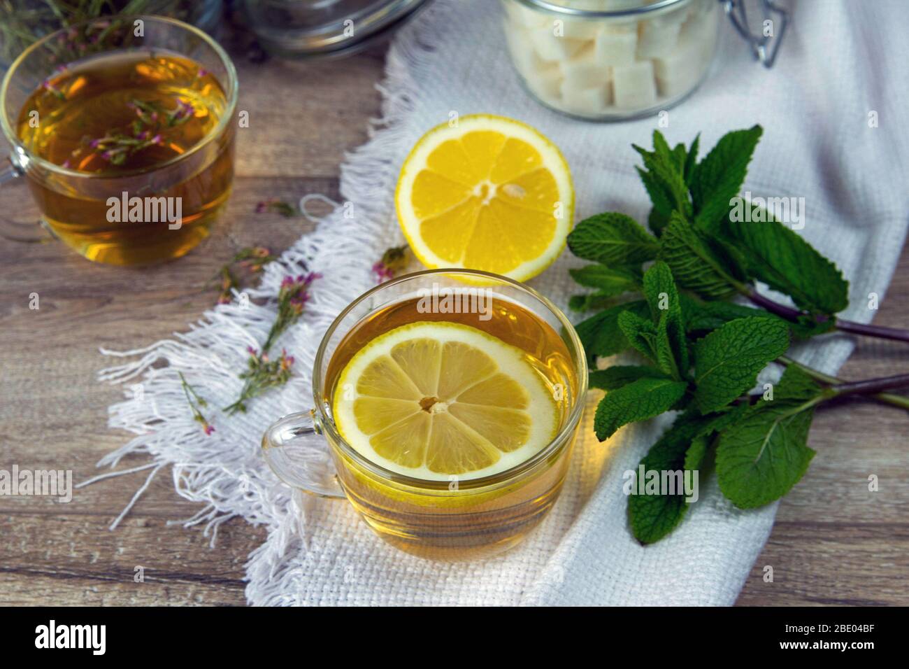 Tasse transparente en verre avec support à thé à la menthe sur un support en bois, à côté d'un bol à sucre avec des cubes de sucre, des feuilles de citron et de menthe tranchées Banque D'Images