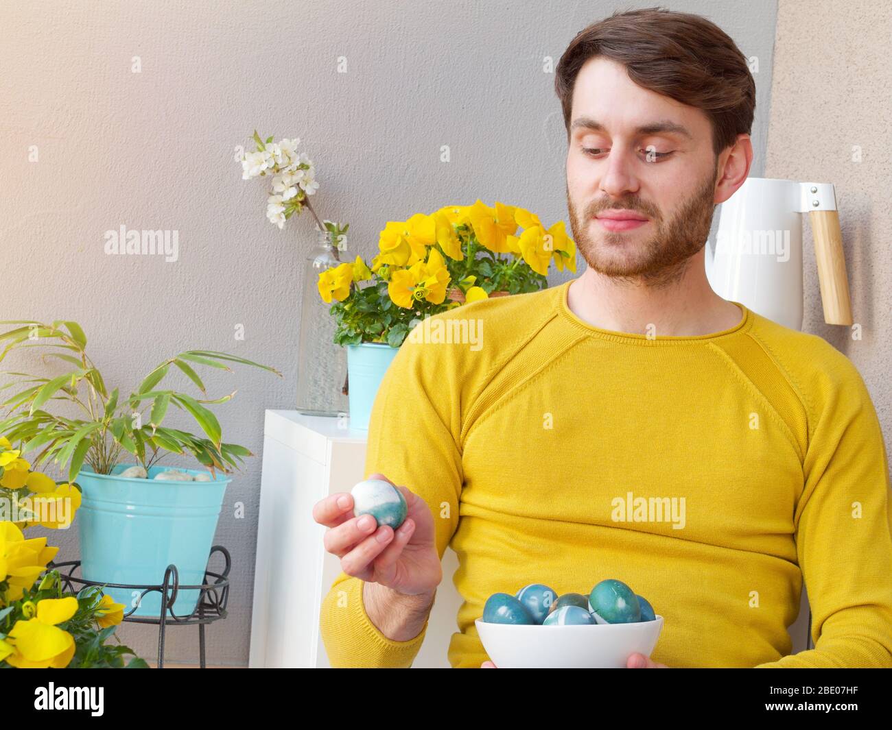 Homme caucasien tenant un bleu des oeufs de pâques dans un bol pendant les temps de pâques, coloré avec la coloration naturelle du chou rouge, portant une bruyère jaune. Banque D'Images
