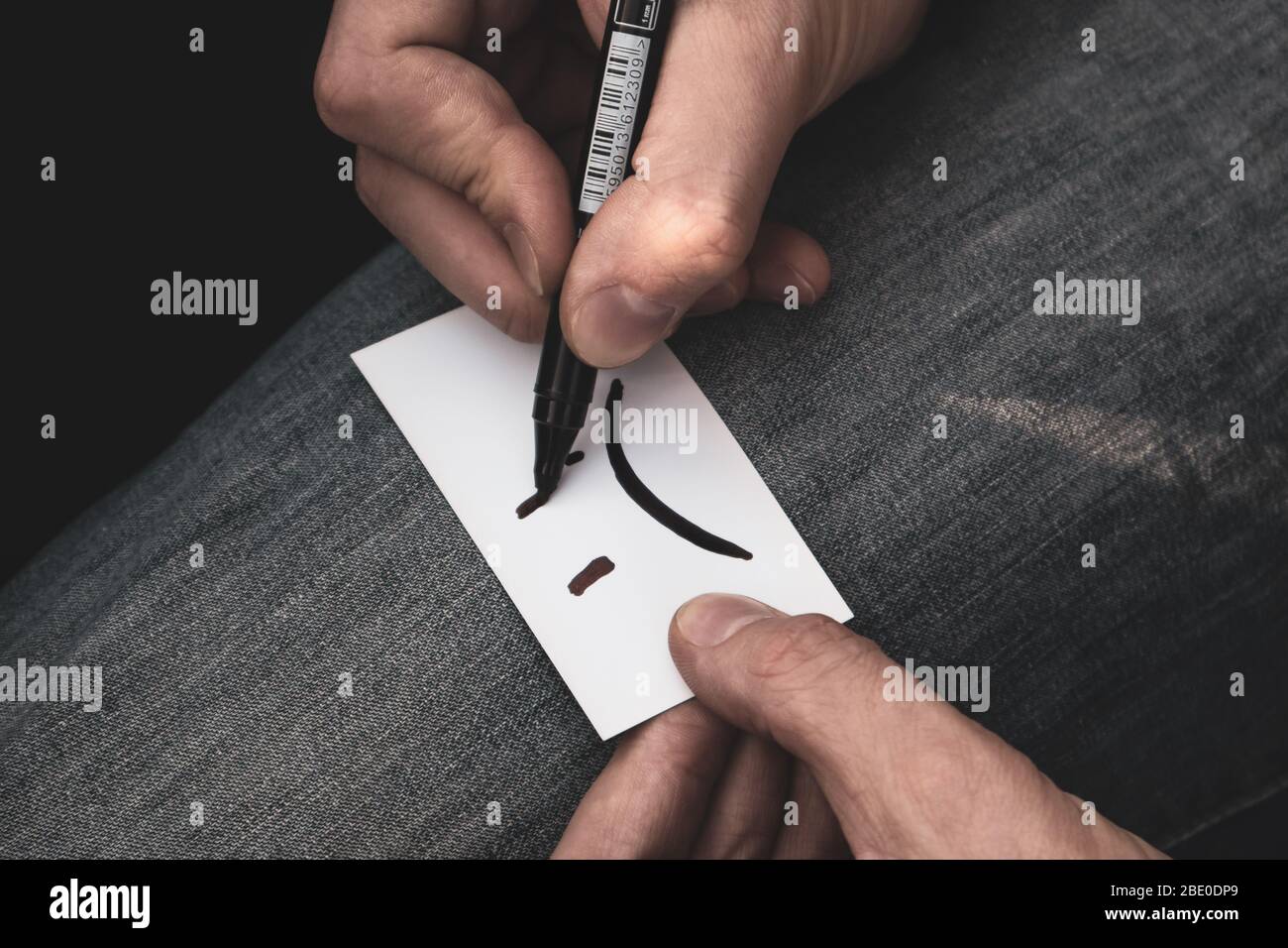 Écrire un émotiji sourire très triste sur la carte de papier blanc dans les mains sur le jeans texture couleur fond dégradé. Triste humeur mauvais temps incolore Banque D'Images