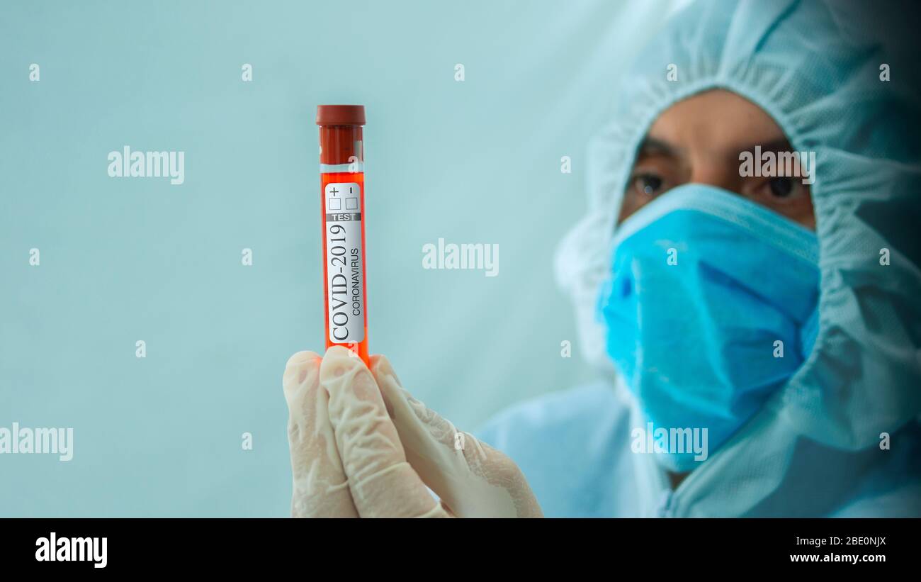 Médecin avec masque et costume blanc de bioprotection tenant un tube de test d'échantillon de sang étiqueté Covid-19 avec sa main et en frappant avec soin sur le dos blanc Banque D'Images