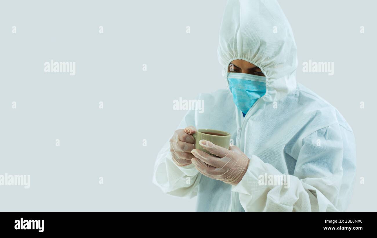 Médecin avec masque bleu et costume bioprotecteur tenant une tasse de café verte avec ses mains, avec une attitude de fatigue, d'inquiétude ou de tristesse sur le ba blanc Banque D'Images