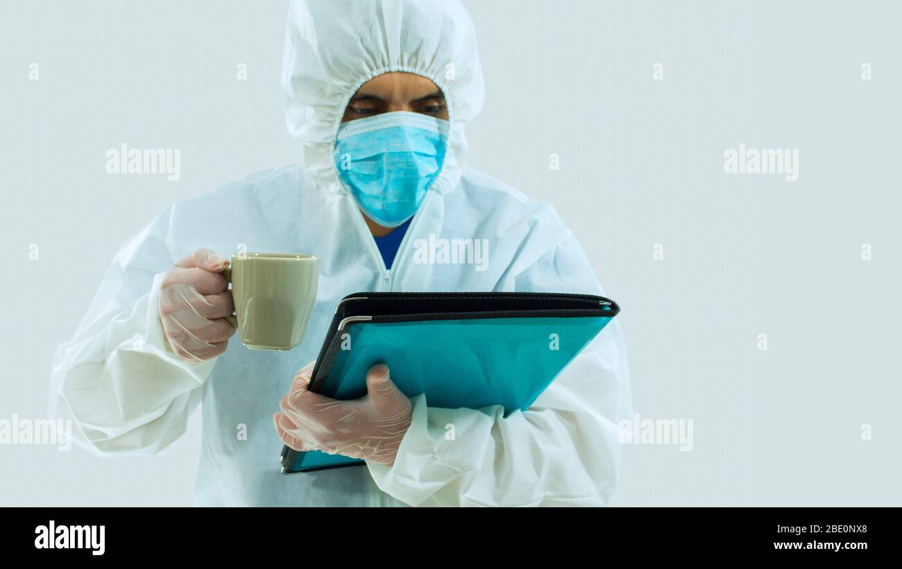 Médecin avec masque bleu et costume bioprotecteur lecture d'un dossier qu'il tient et une tasse de café verte dans sa main droite sur fond blanc Banque D'Images