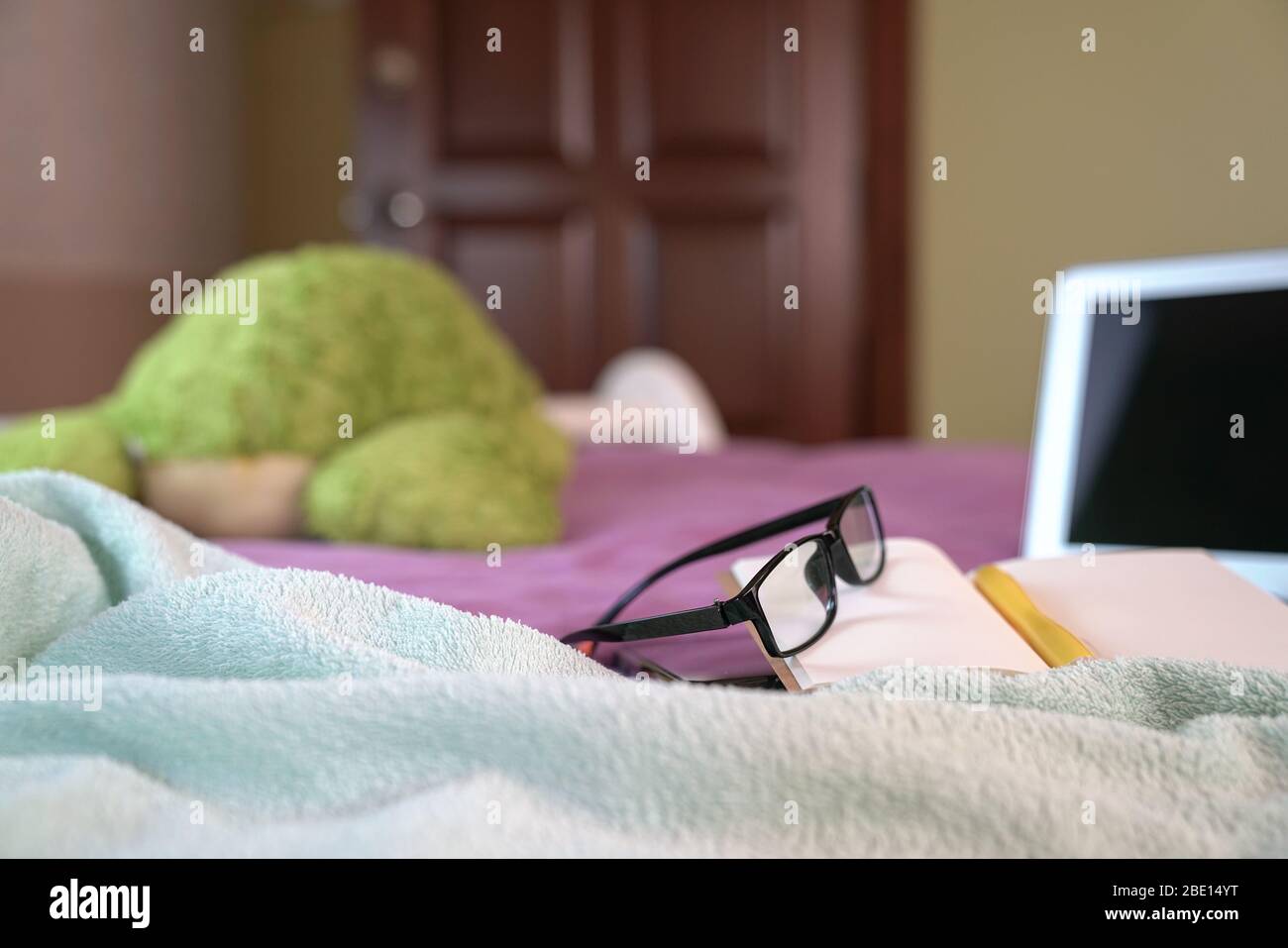 Ordinateur portable avec bloc-notes, stylo, lunettes de lecture et jouet doux sur le dessus du lit dans la chambre. Travailler à la maison. Se concentrer sur les lunettes. Banque D'Images