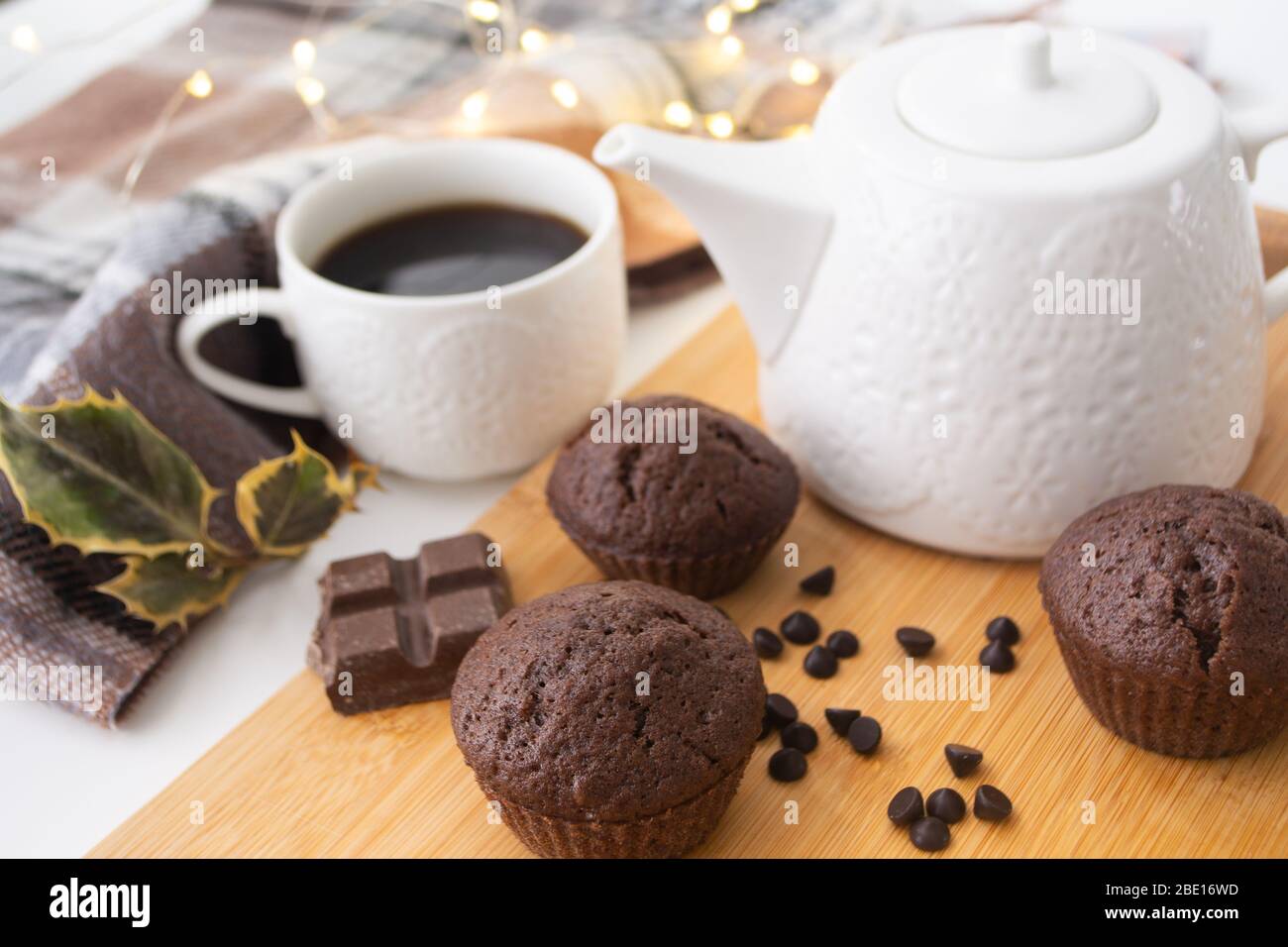 Muffins au chocolat, chips au chocolat, bar au chocolat, tasse de café, théière sur une surface en bois avec écharpe chaude, lumières de Noël bokeh et houx dans le Banque D'Images