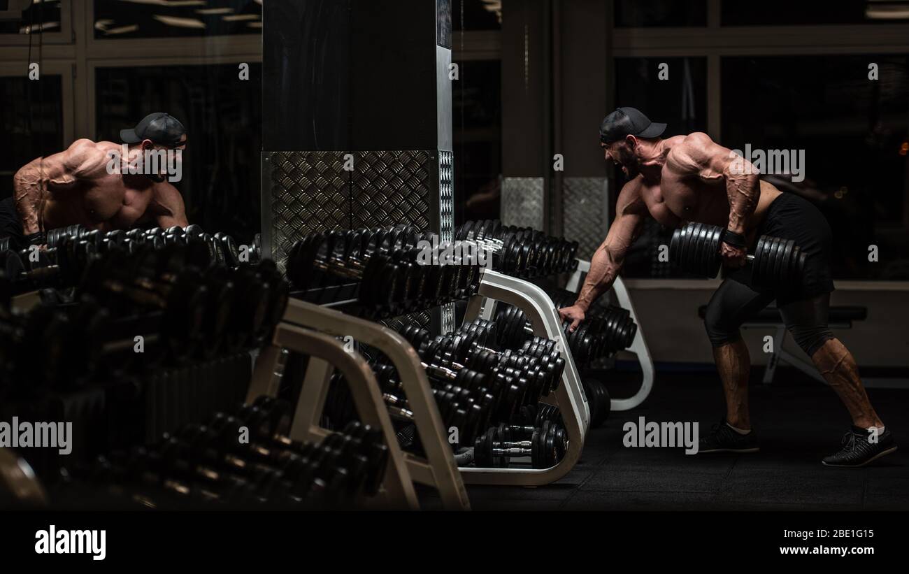 jeune homme caucasien barbu dans le chapeau et les malles de sport soulevant poids lourd haltère dans la nuit sombre salle de fitness Banque D'Images