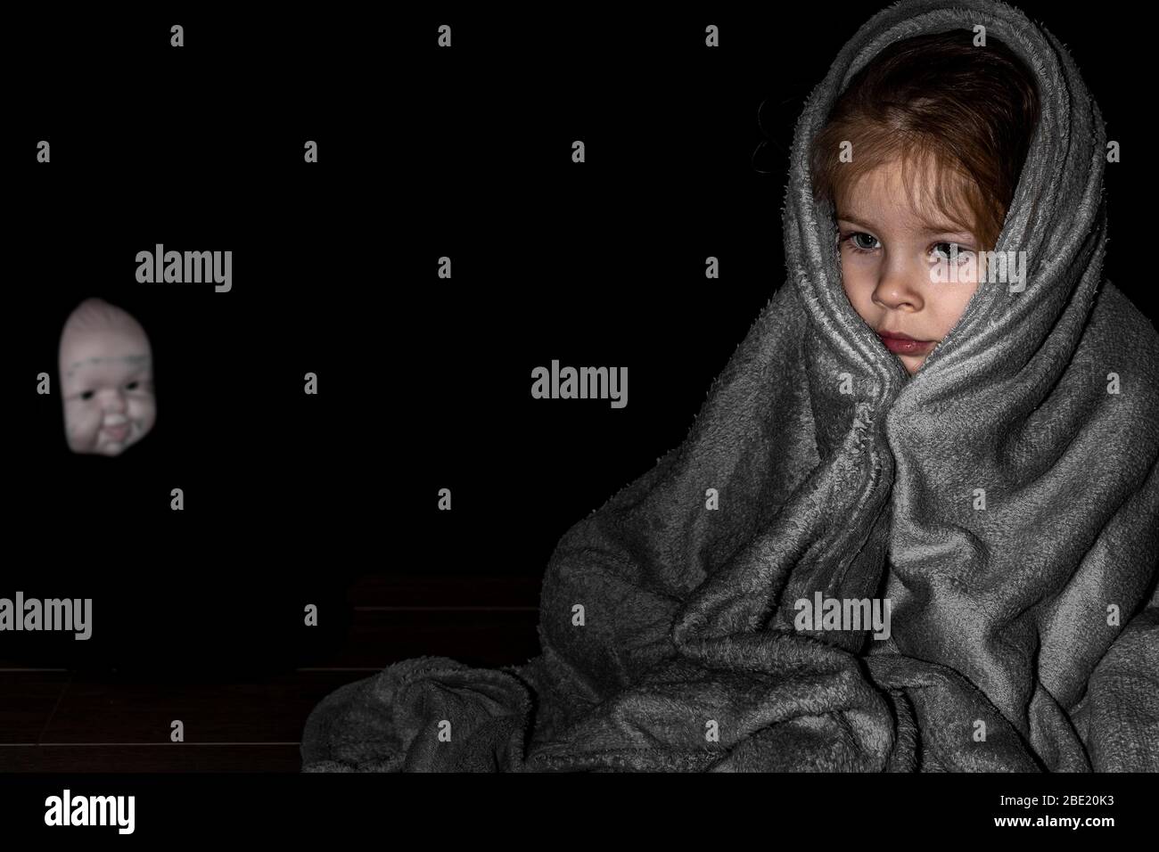 une petite fille assise dans le noir enveloppé dans une couverture, à l'arrière-plan le visage flou d'une poupée effrayante. la nuit des enfants craint Banque D'Images