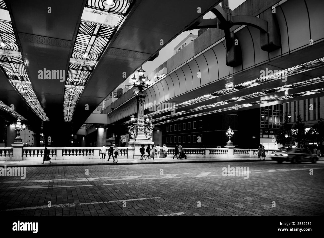 Quatre lampes en forme de kirin brillent d'une lueur dorée sous l'autoroute surélevée du pont de Nihonbashi à Tokyo. Un taxi aux feux arrière rouge vif traverse l'intersection. Des piétons traversent le pont en arrière-plan. Achevé en 1911, le pont sert de point zéro pour toutes les distances au Japon. Banque D'Images