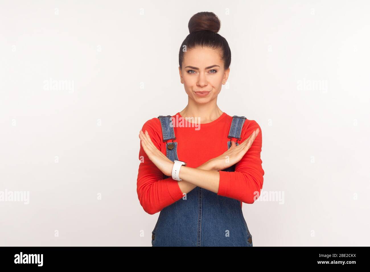 Pas d'accès ! Portrait d'une fille sérieuse et stricte avec un pain de cheveux dans des salopettes en denim montrant un signe d'arrêt avec des mains croisées, rejetant l'offre, l'expression d'avertissement. Banque D'Images