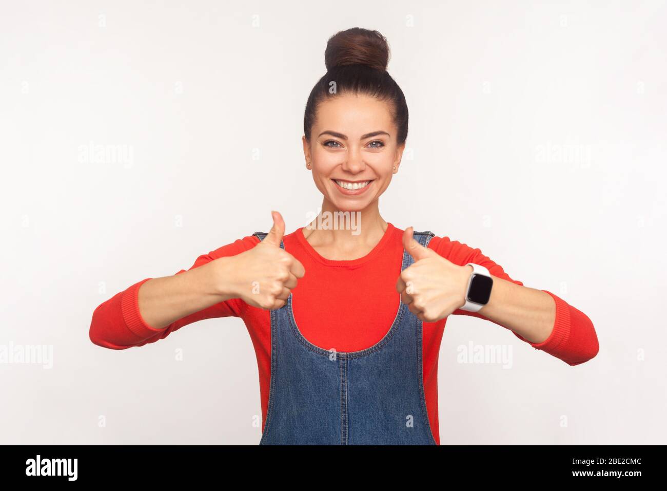 Excellent travail ! Portrait d'une jolie fille optimiste et élégante avec un pain à cheveux dans des salopettes en denim montrant des pouces vers le haut, montrant comme, geste d'approbation pour les succes Banque D'Images