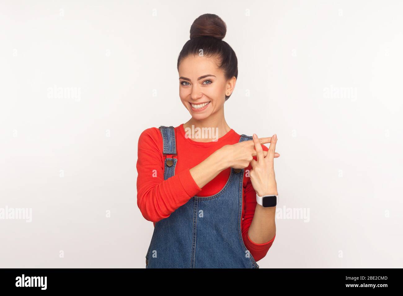 Portrait d'une fille optimiste et heureuse avec un pain de cheveux dans des salopettes en denim montrant le geste de hashtag et sourire à l'appareil photo, symbole du contenu viral, blog Banque D'Images