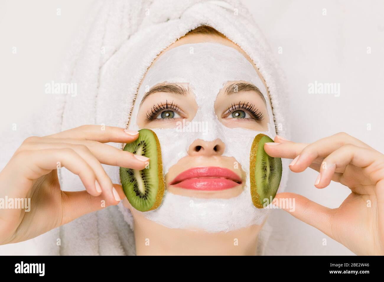 Gros plan sur une jolie femme souriante avec masque hydratant blanc et morceaux de kiwi frais sur les joues. Masque facial de beauté, soins de la peau et traitement Banque D'Images