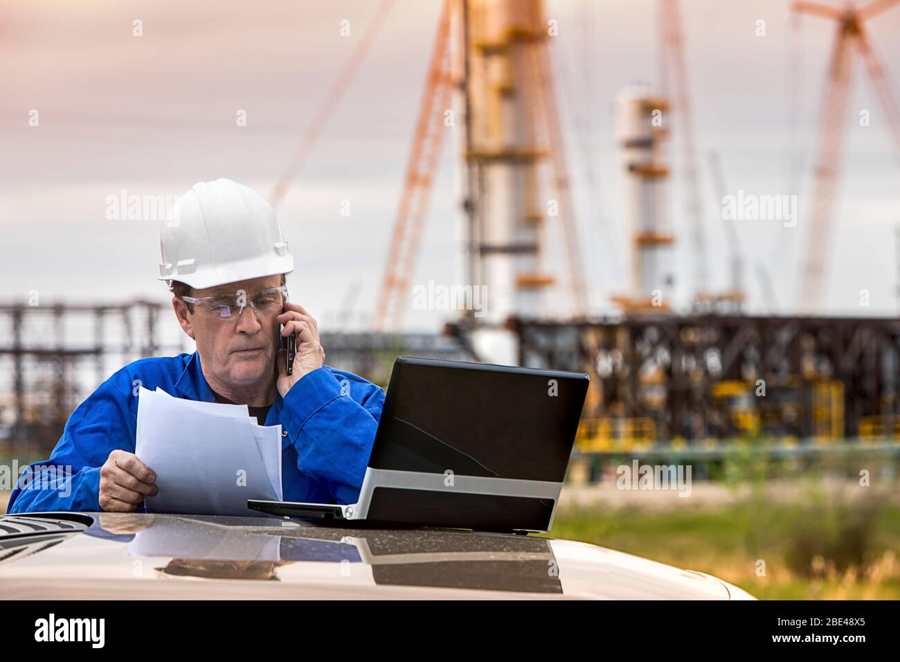 L'homme travaille sur un ordinateur portable et un smartphone tout en se tenant à un véhicule avec une plate-forme pétrolière en arrière-plan; Alberta, Canada Banque D'Images