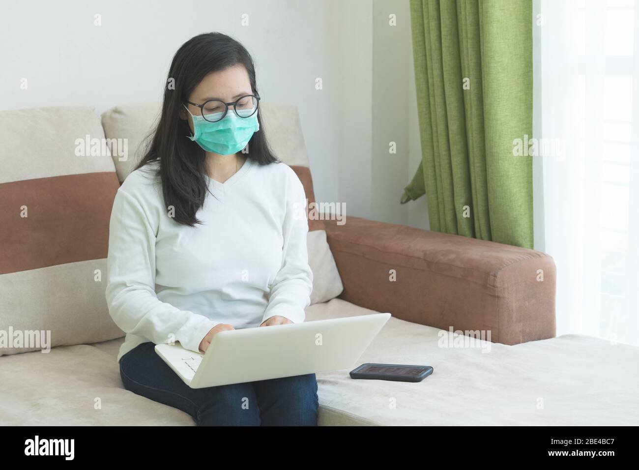 Jeune femme belle waring masque médical assis sur le canapé et télétravailler sur son ordinateur portable dans la chambre blanche. Travaillez à domicile pour un conceps social distancer Banque D'Images