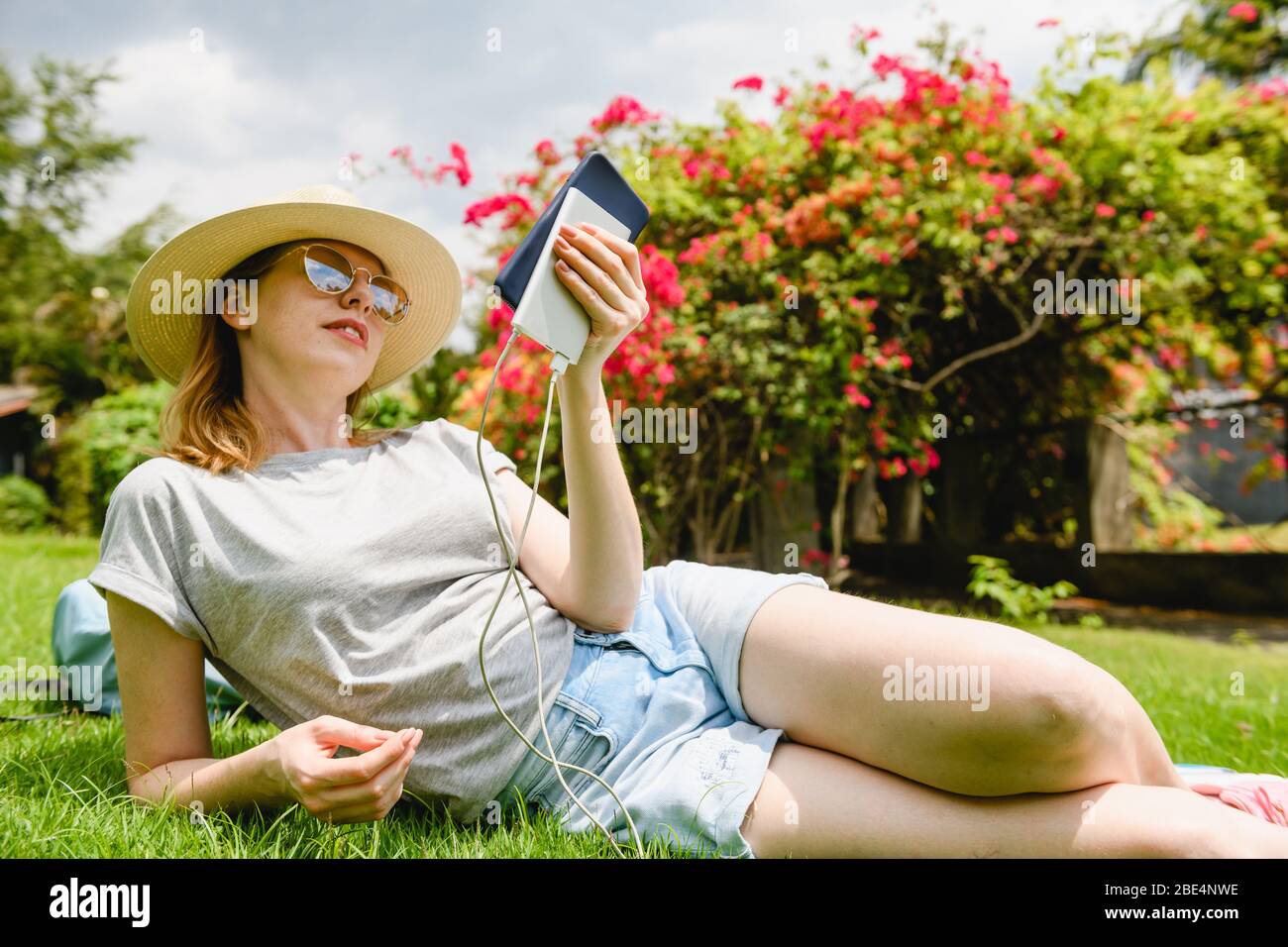 Une femme s'est couponée sur l'herbe verte, tenant le téléphone mobile et la banque d'alimentation portable au pique-nique dans le parc Banque D'Images