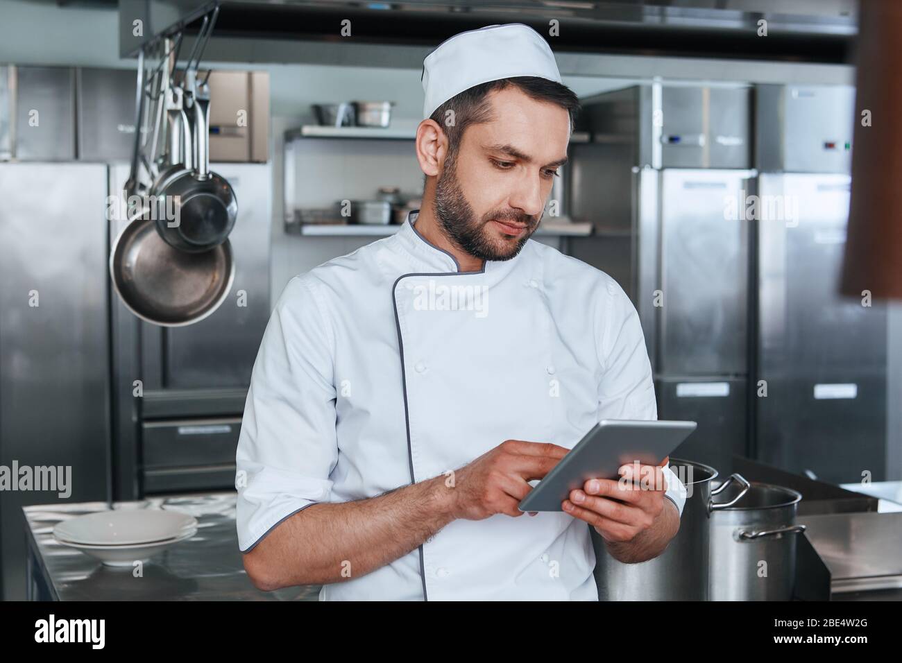 Le chef barbu mâle regarde une tablette numérique tout en préparant la nourriture dans la cuisine du restaurant Banque D'Images