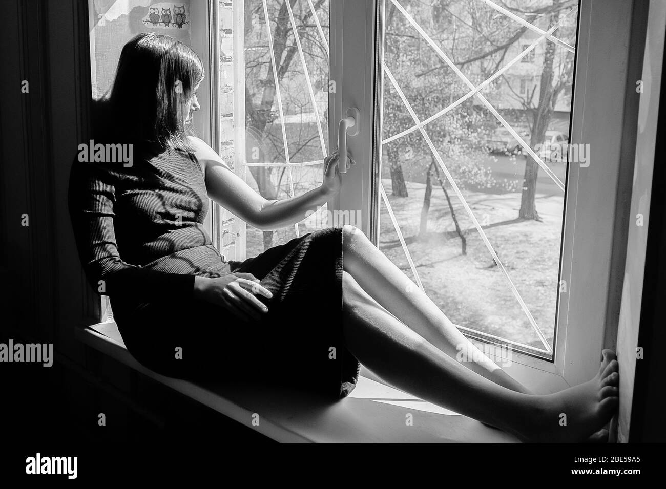 portrait noir et blanc d'une belle fille à poil noir assise sur un seuil de fenêtre dans une robe sombre bien ajustée Banque D'Images