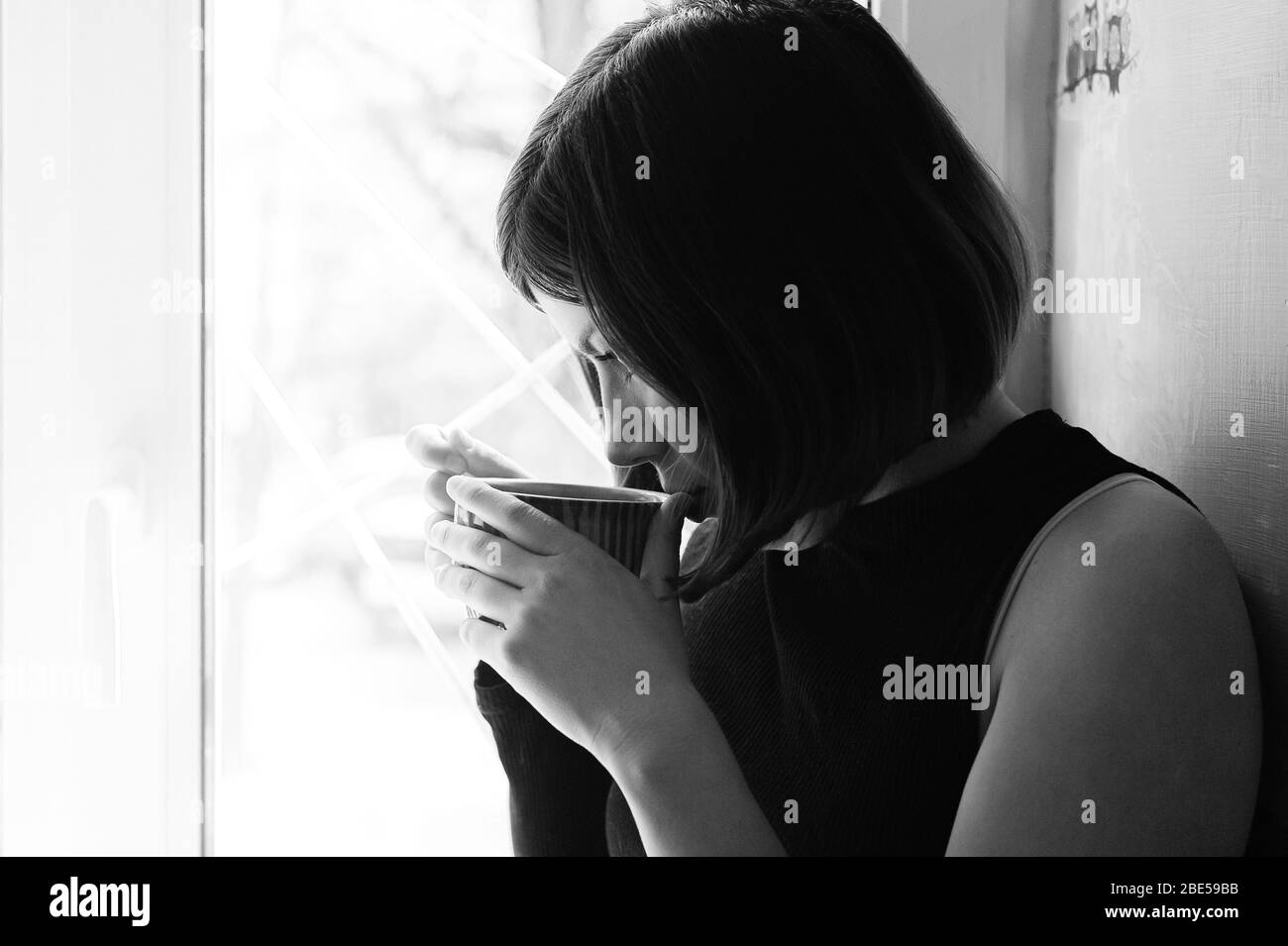 portrait noir et blanc d'une jeune fille de brunette en noir avec une grande tasse de thé par la fenêtre Banque D'Images