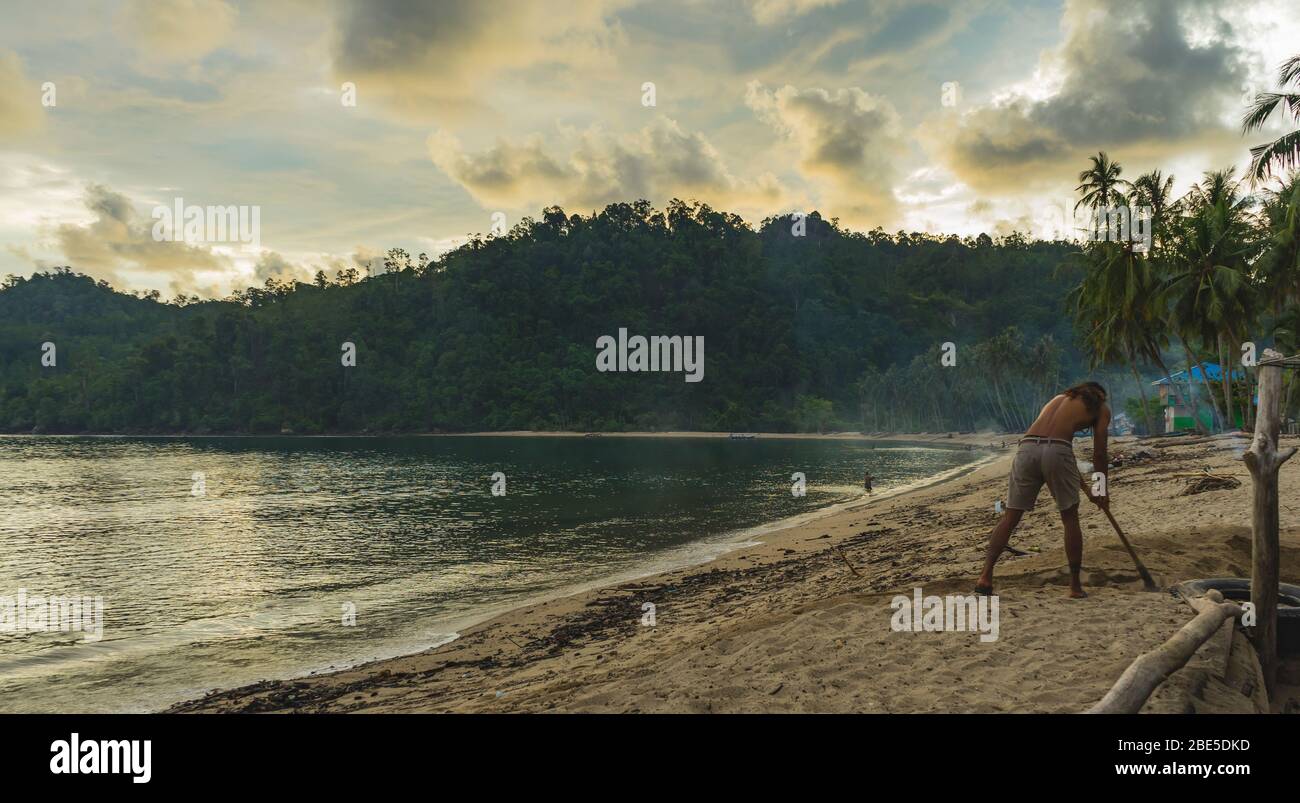 03 juillet 2018 Sungai Pinang, Sumatra, Indonésie : scène tranquille de village de pêcheurs avec un homme local taté creusant du sable. Palmiers et montagnes autour Banque D'Images
