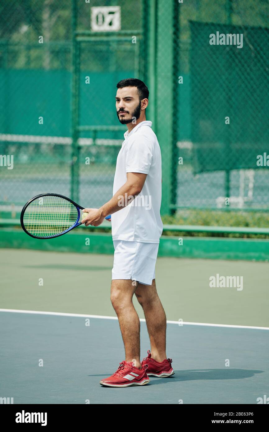 Beau jeune homme hispanique dans des vêtements de sport blancs jouant au tennis à l'extérieur Banque D'Images