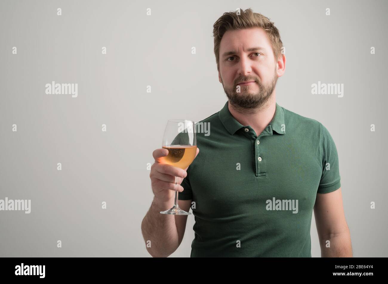 Portrait d'un homme élégant et sérieux avec une barbe épaisse, vêtu d'un t-shirt vert décontracté, qui tient le verre de vin à l'air d'un appareil photo isolé sur le gris Banque D'Images