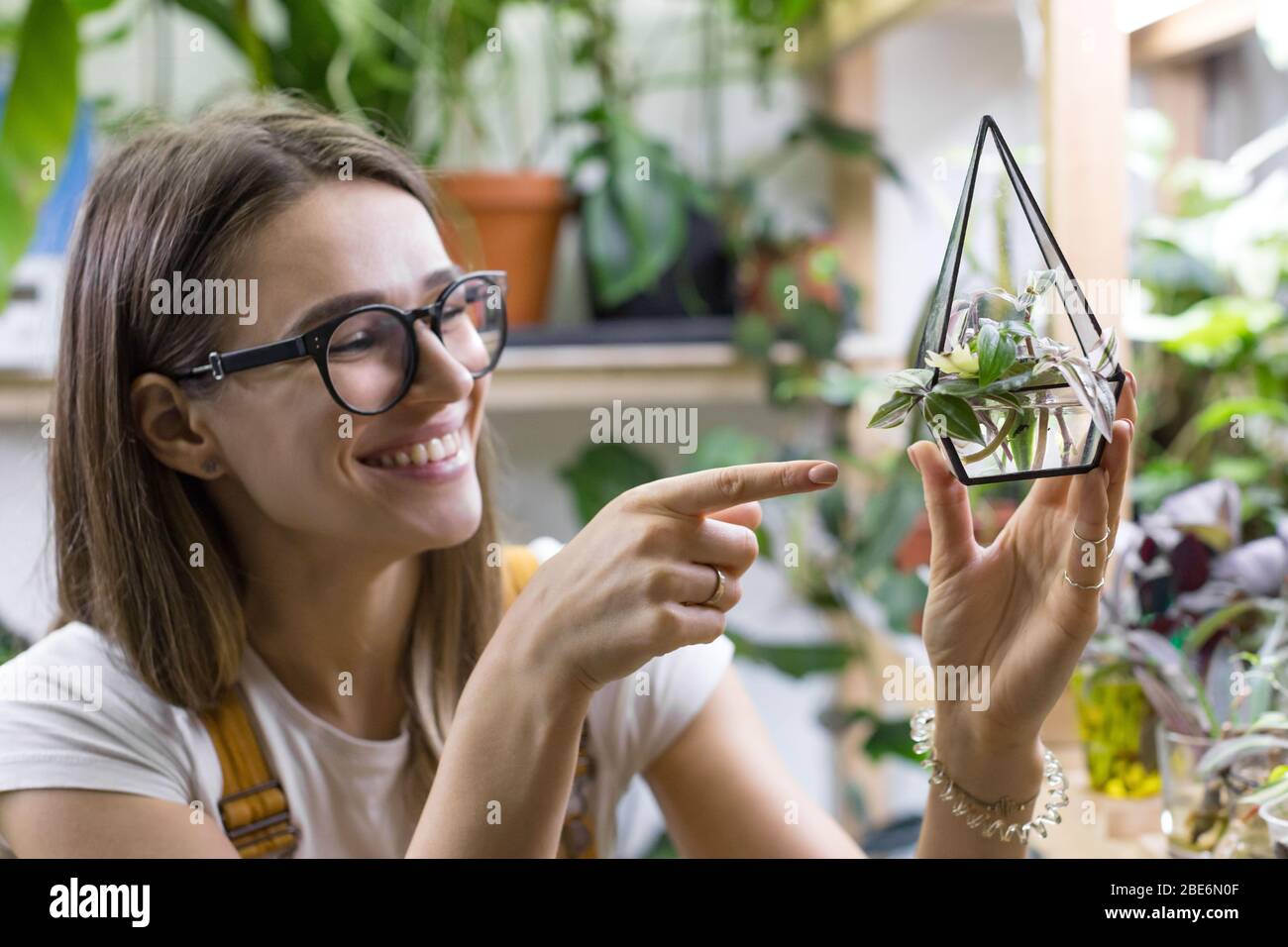 Le jardinier souriant de femmes dans les lunettes montre index sur les boutures de tradescantia dans un petit florarium de verre pour la germination des plantes, potted houseplan Banque D'Images