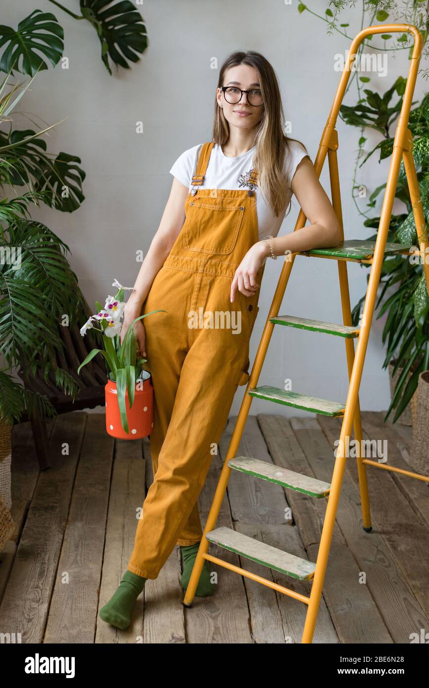 Jeune femme jardinier dans des verres portant des combinaisons, tenant l'orchidée dans la vieille carafe à lait rouge et se penchant sur l'échelle orange vintage, regardant l'appareil photo. Accueil Banque D'Images