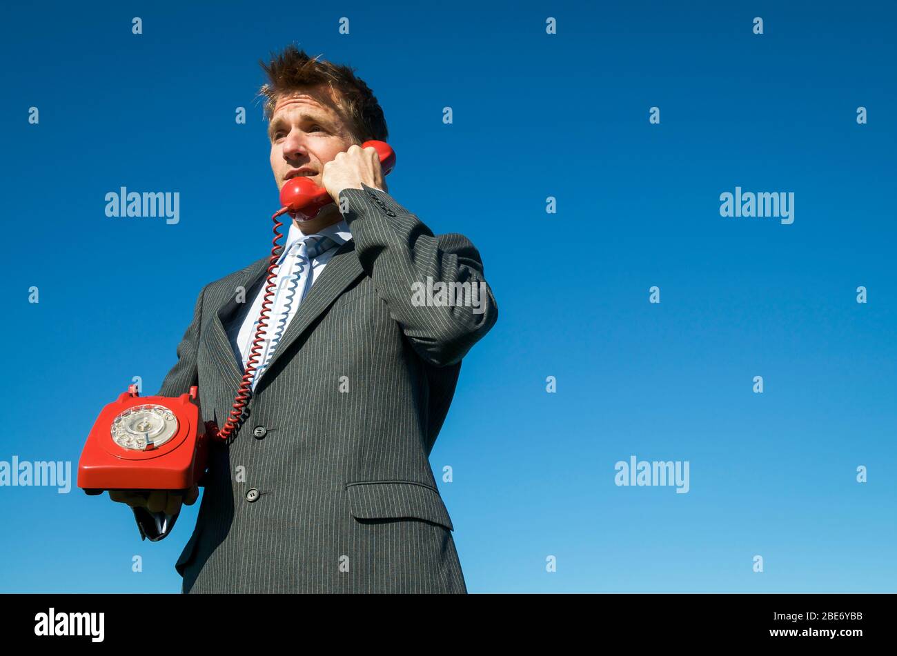 Homme d'affaires ayant une conversation urgente sur un téléphone rotatif rouge à l'ancienne debout à l'extérieur dans le ciel bleu ensoleillé Banque D'Images