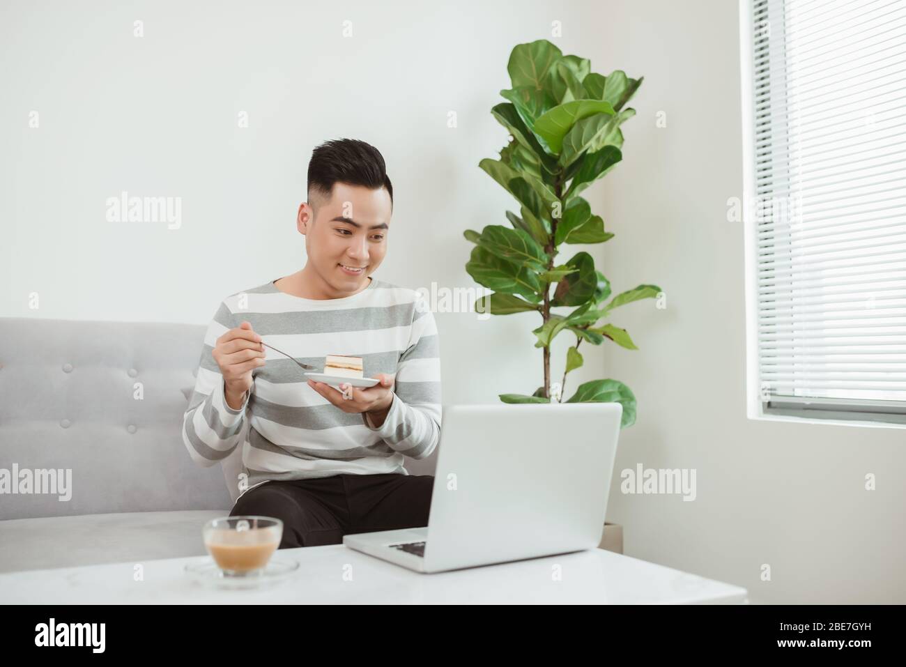 Jeune homme asiatique mangeant des gâteaux lorsqu'il est assis sur un canapé et à l'aide d'un ordinateur portable. Travailler à domicile. Banque D'Images