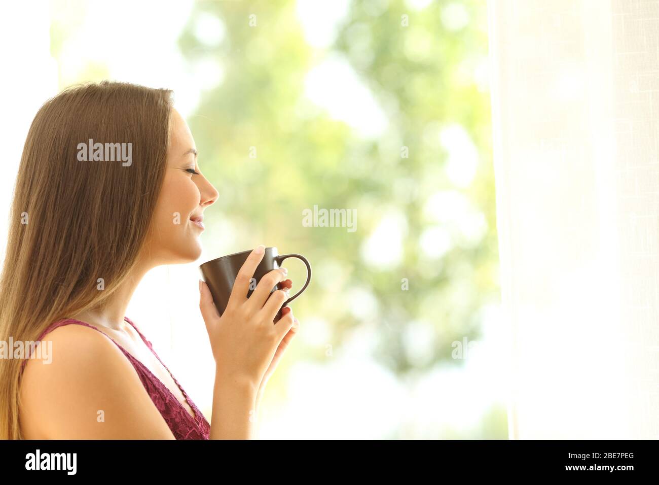 Portrait de la vue latérale d'une femme satisfaite qui sent une tasse de café debout à côté de la fenêtre à la maison Banque D'Images